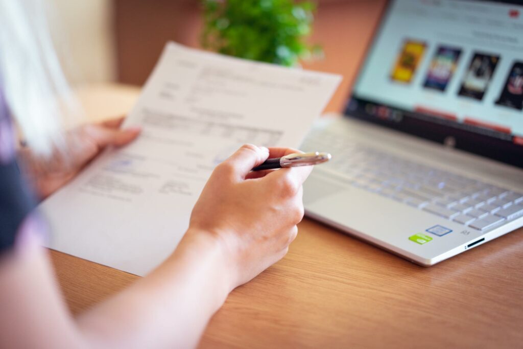 A person holding a pen reviews a printed document at a wooden desk. A laptop with a blurred webpage is open nearby on the right. A small green plant is visible in the background.