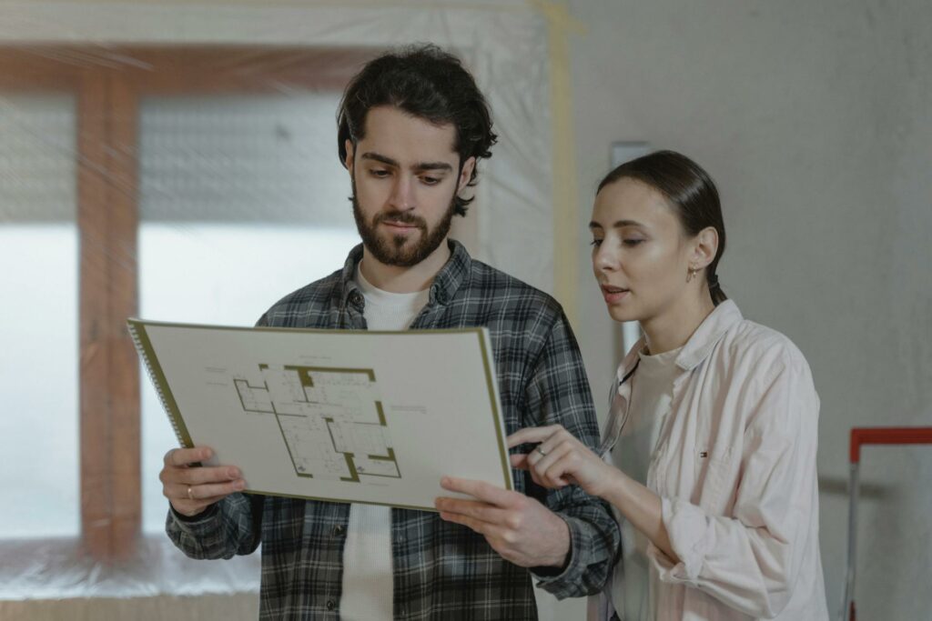 A man and a woman examine a floor plan in a room under renovation. The man, wearing a plaid shirt, holds the plan while the woman, in a light jacket, points at it. A window covered with plastic is in the background.